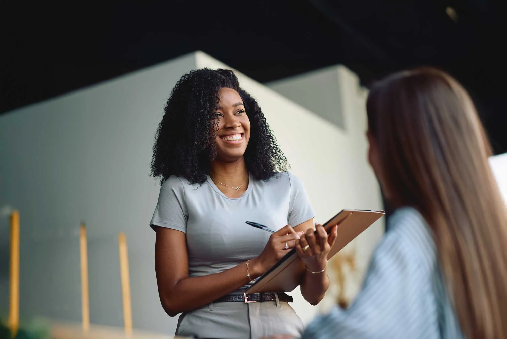 Smiling woman holding clipboard and pen, speaking with another person with long hair.