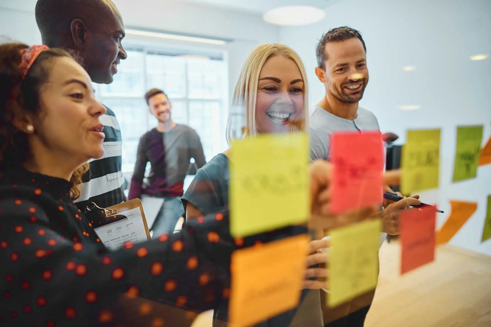 Five diverse coworkers smiling and discussing colorful sticky notes on a glass wall in an office.