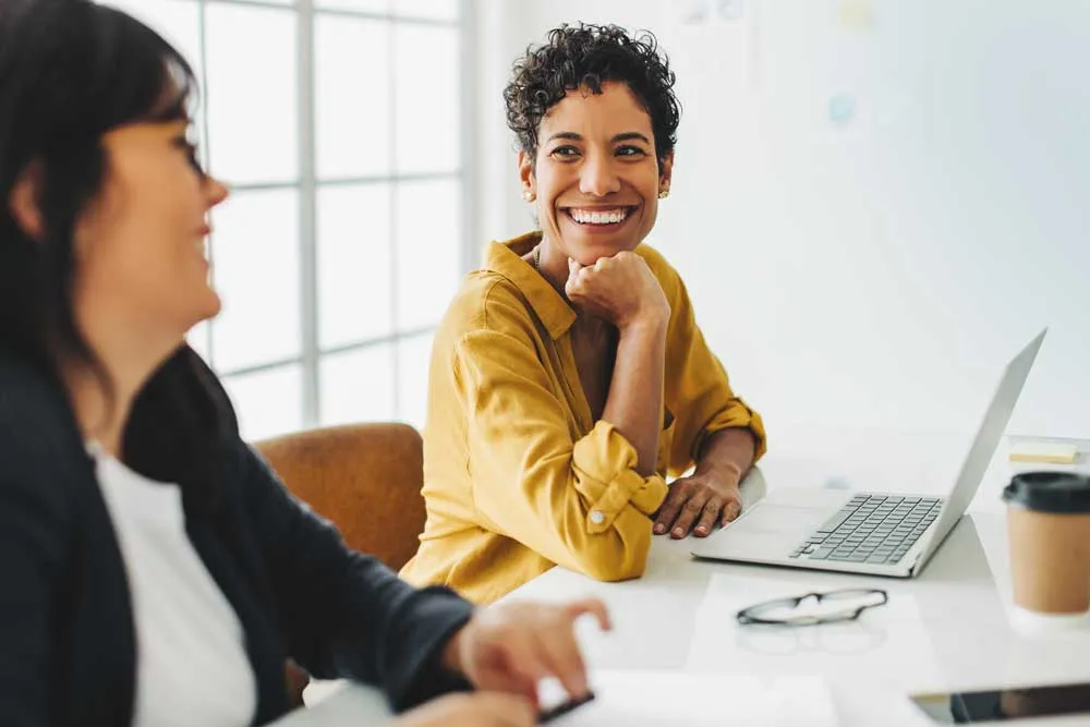 Two women sitting at a desk, one smiling while looking at the other, with a laptop and coffee cup present.