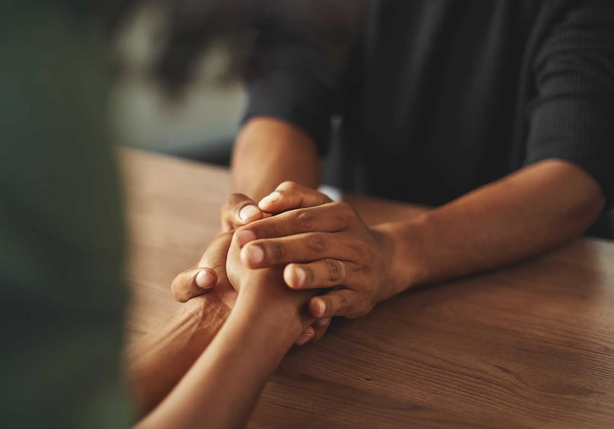 Two people sitting at a wooden table holding hands in a gesture of support and comfort.