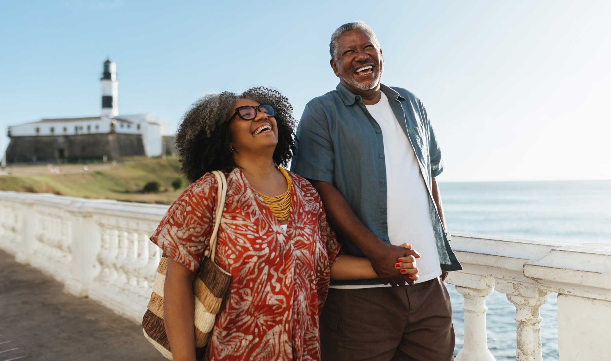 Smiling couple holding hands outdoors near a white balustrade by the sea with a lighthouse in the background.