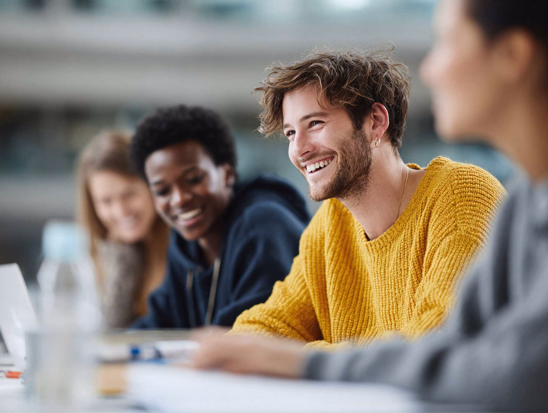 Young diverse people sitting at a table smiling and talking, with one person in a yellow sweater.