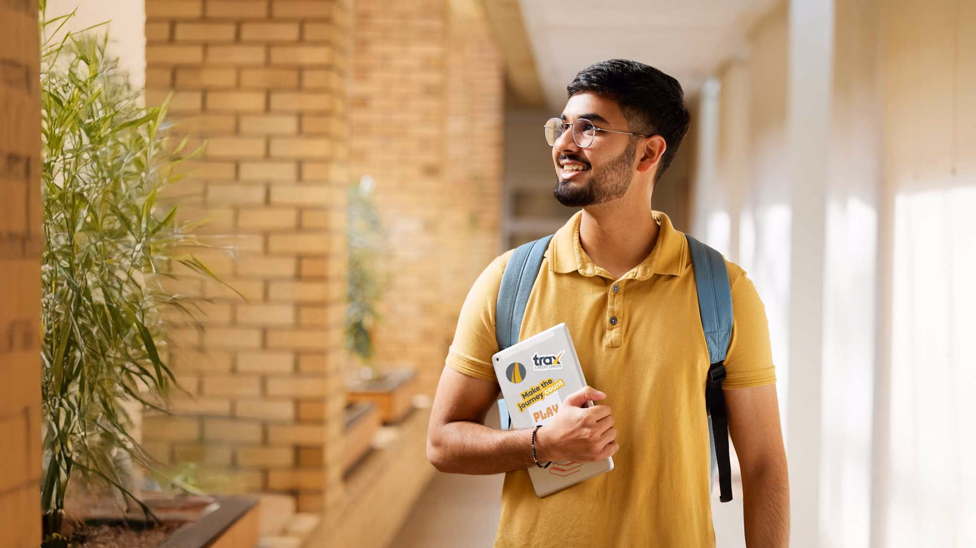 Smiling young man in yellow shirt holding tablet and wearing a blue backpack in a bright hallway.