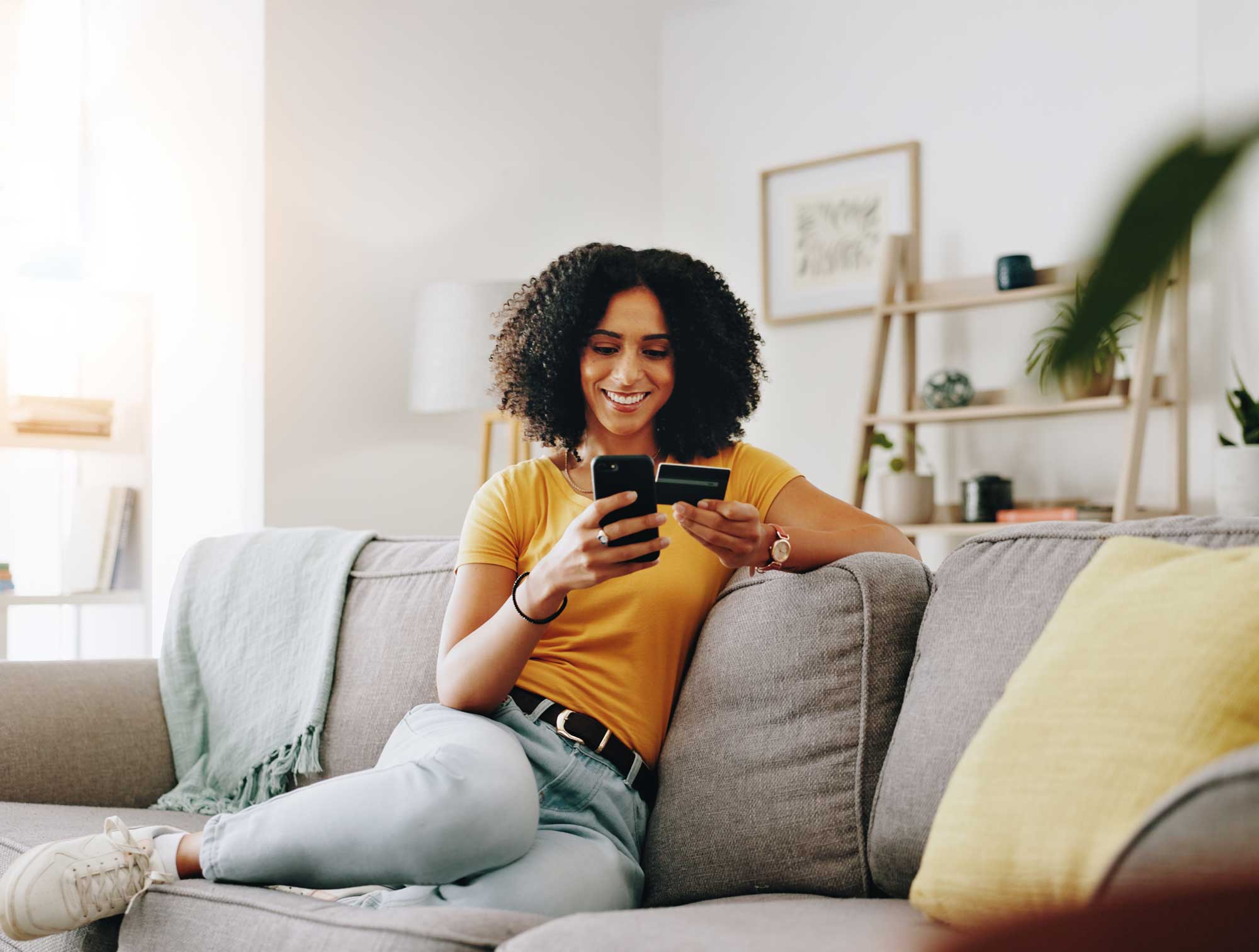 Young woman sitting on a gray couch in a bright living room, smiling while holding a smartphone in one hand and a credit card in the other, appearing to make an online payment.