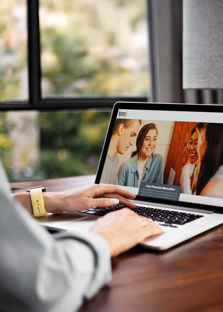 Person using a laptop with a website open showing three smiling people engaged in conversation, titled Trax Financial Education, on a wooden table near a window.