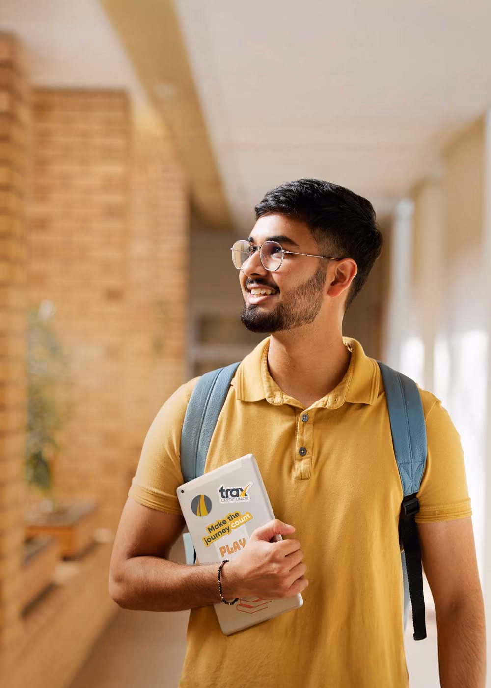 Smiling young man in yellow shirt holding tablet and wearing a blue backpack in a bright hallway.