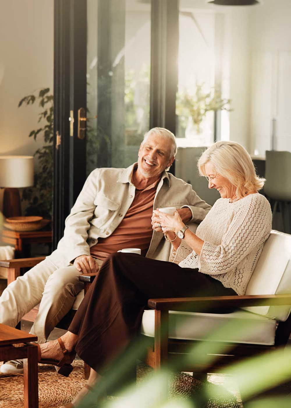 Smiling elderly couple sitting and laughing on a cozy patio with plants around them.