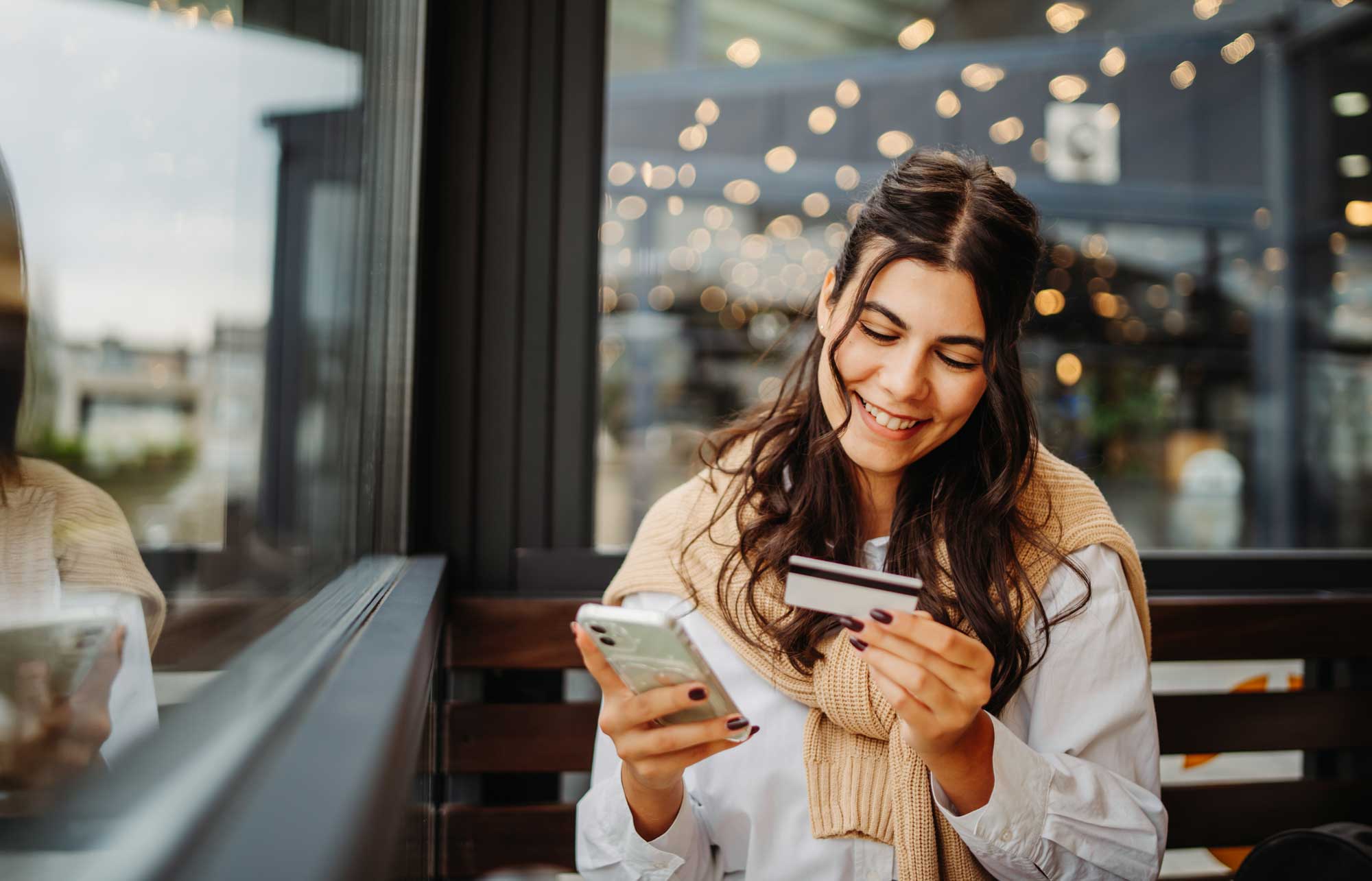 Smiling woman sitting by a window, holding a smartphone in one hand and a credit card in the other, in a cozy indoor setting with blurred lights in the background.