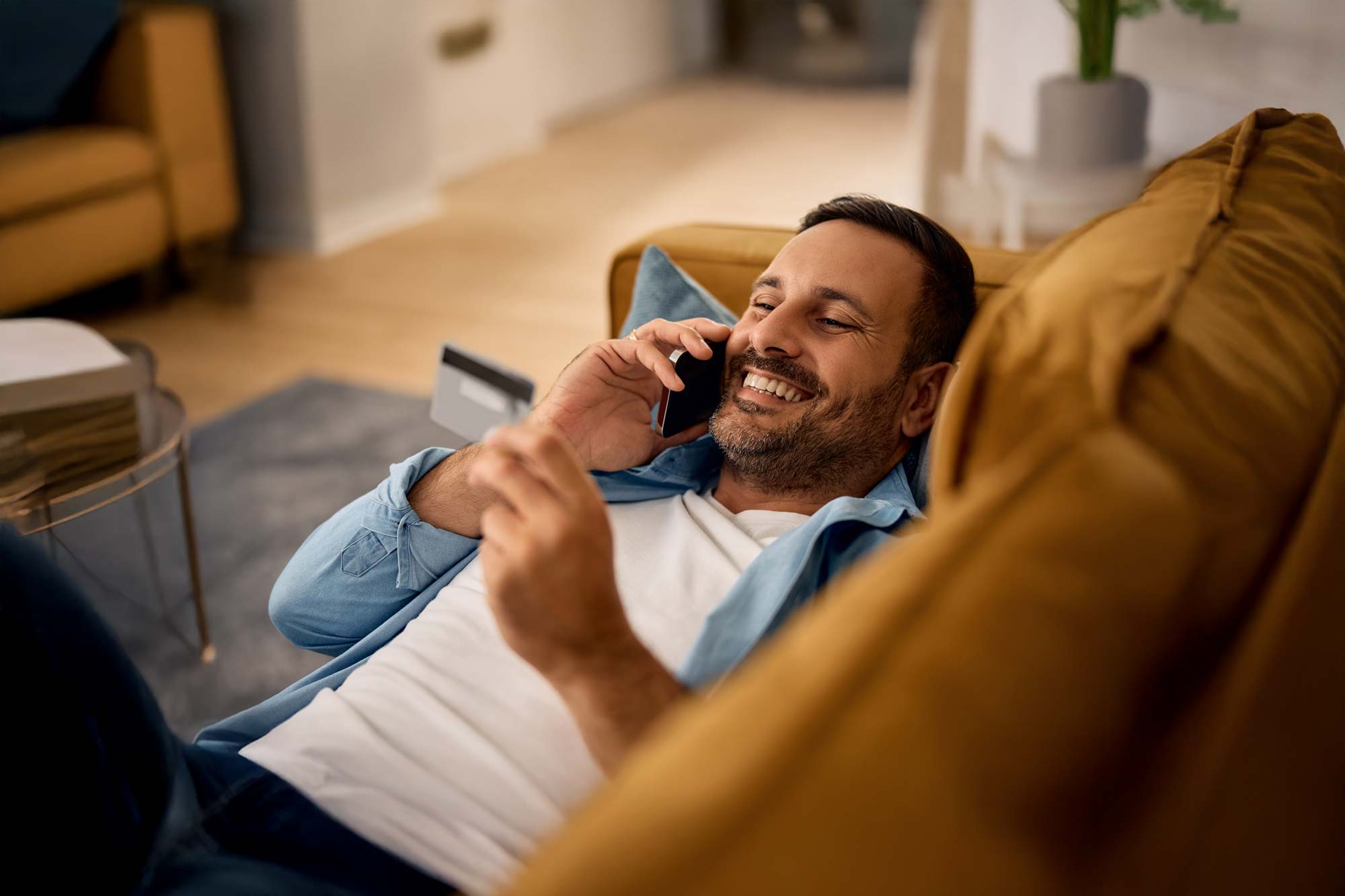 Smiling man lying on a mustard yellow couch talking on a smartphone and holding a credit card in his hand.