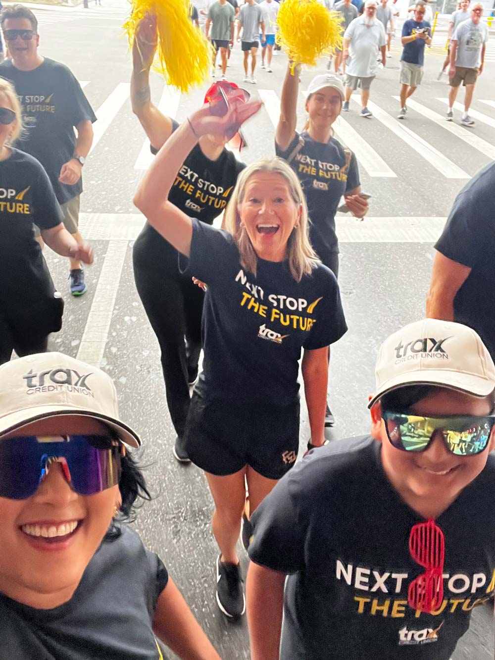A group of smiling people wearing black t-shirts with "Next Stop The Future" and white caps with "trax Credit Union" logo, walking outdoors with two women waving yellow pom-poms.