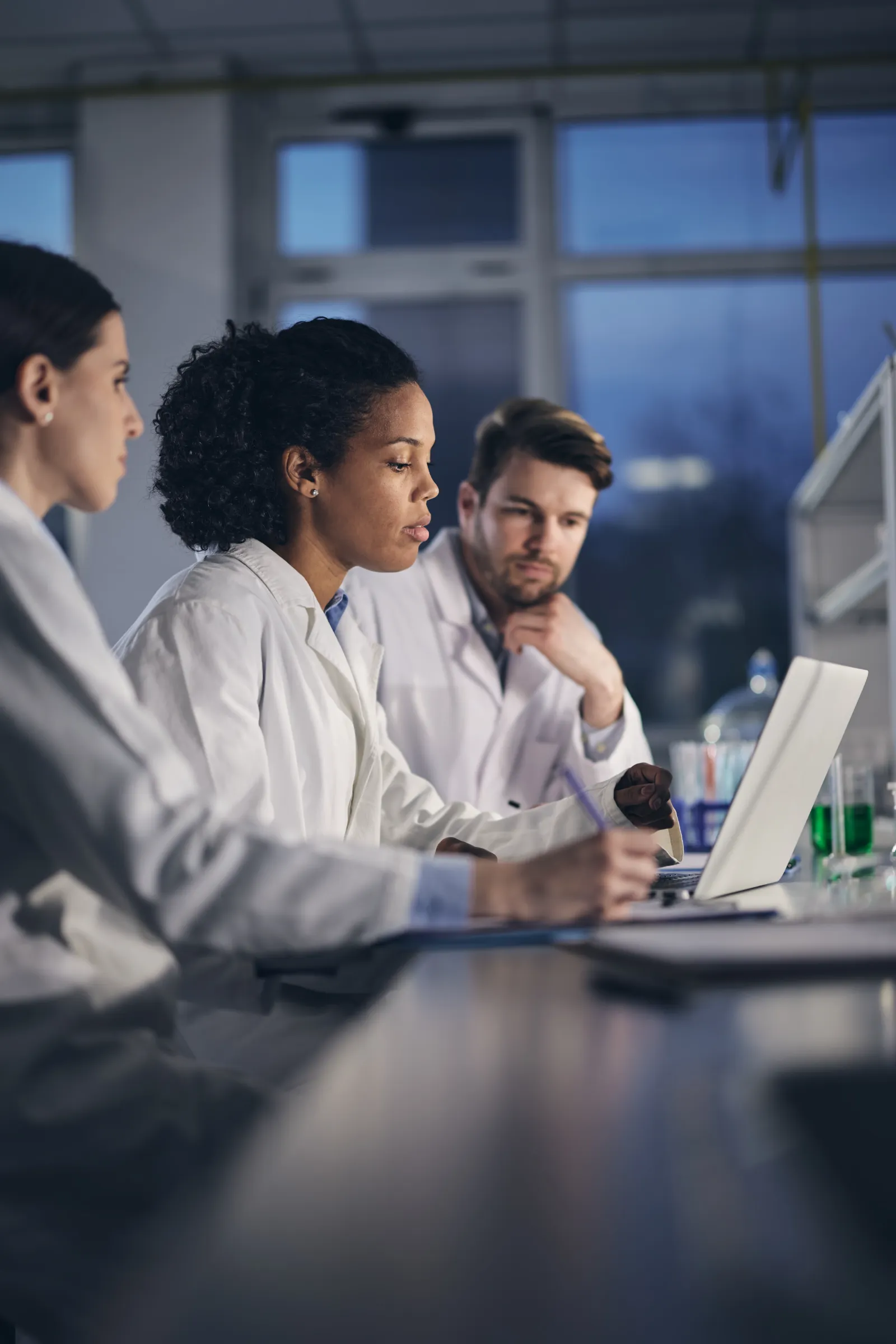 Three researchers in lab coats reviewing data on a laptop in a laboratory