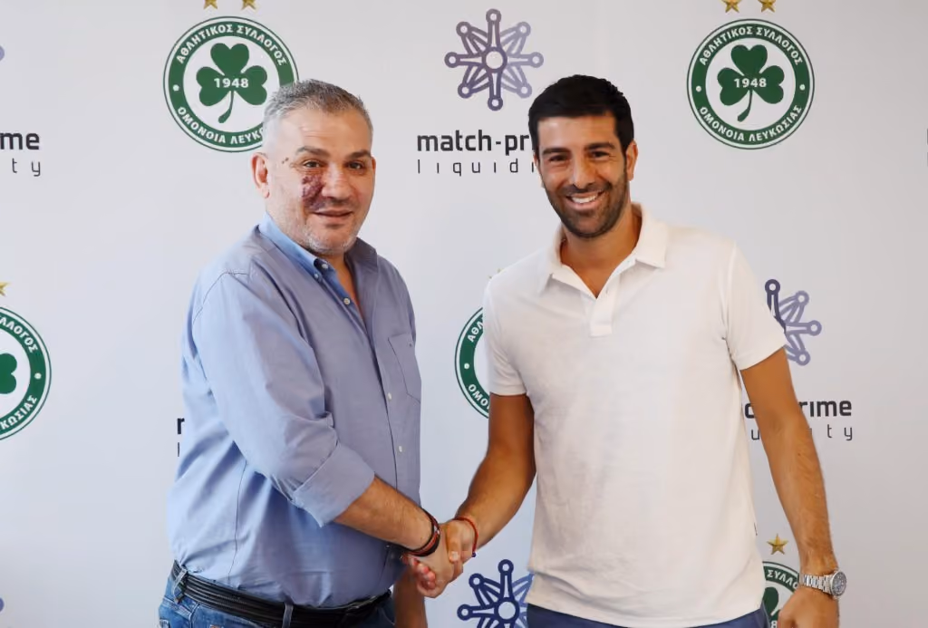 Representatives of Omonoia Nicosia and Match-Prime shaking hands in front of both logos