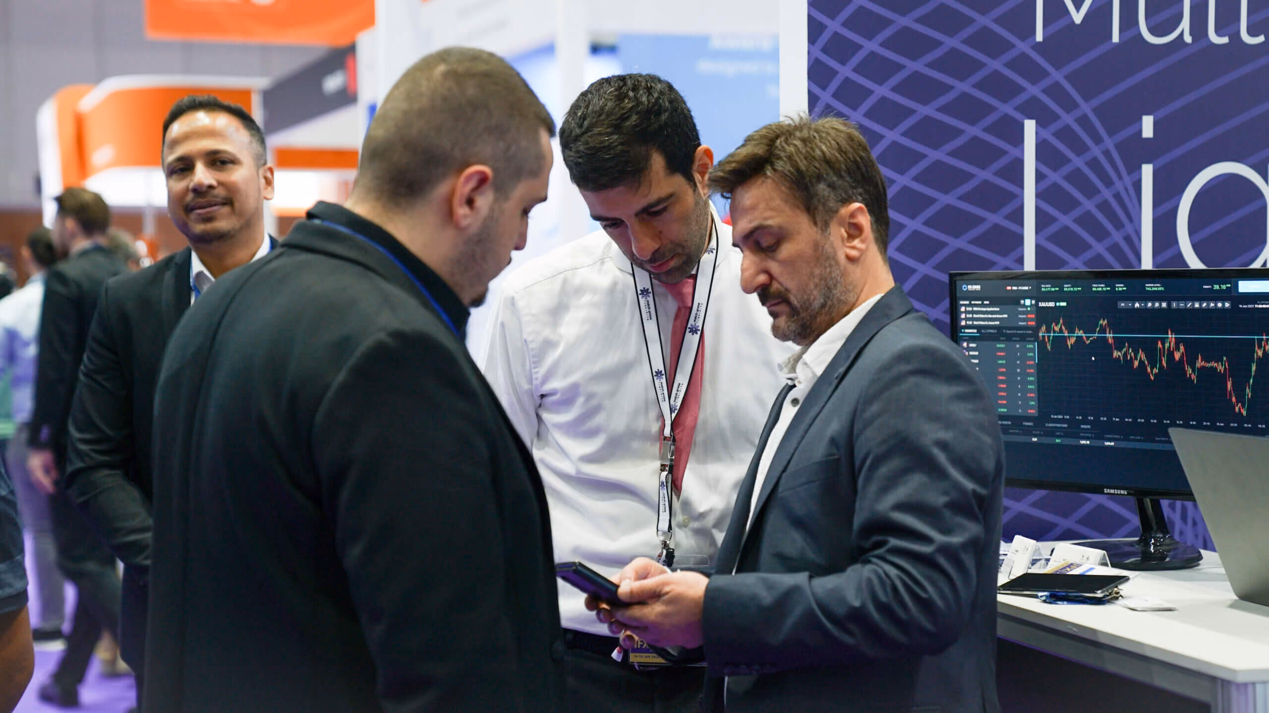 Four attendees in conversation at the Match-Prime stand, beside a monitor showing a live trading chart.