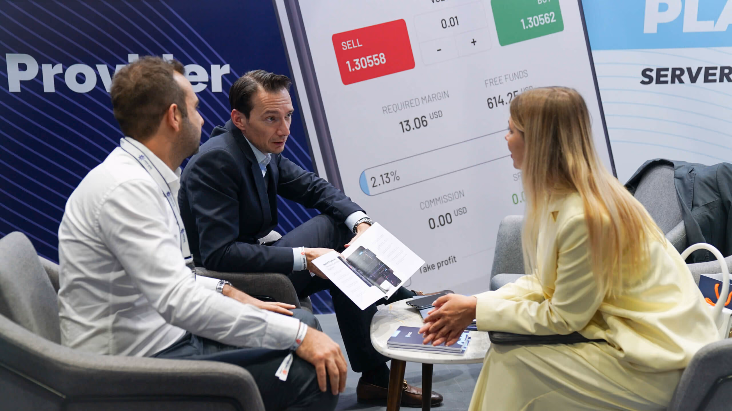Two Match-Prime representatives talking with a visitor in armchairs, a trading platform order ticket on the screen behind them.