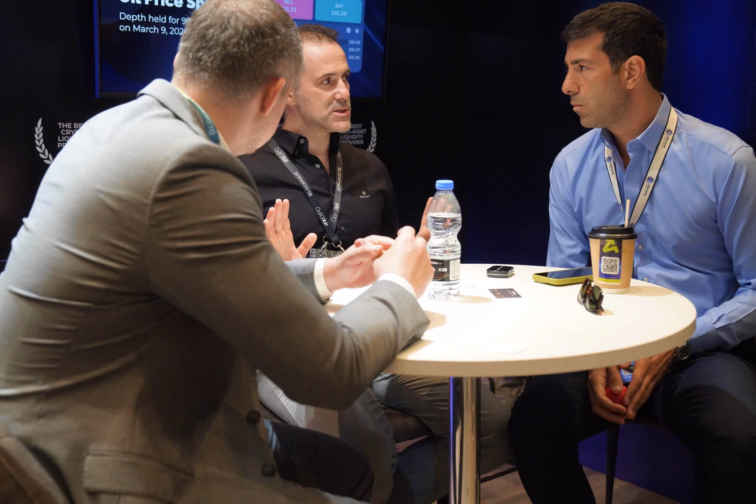 Three men in discussion around a small table at the Match-Prime stand, an execution-data screen behind them.