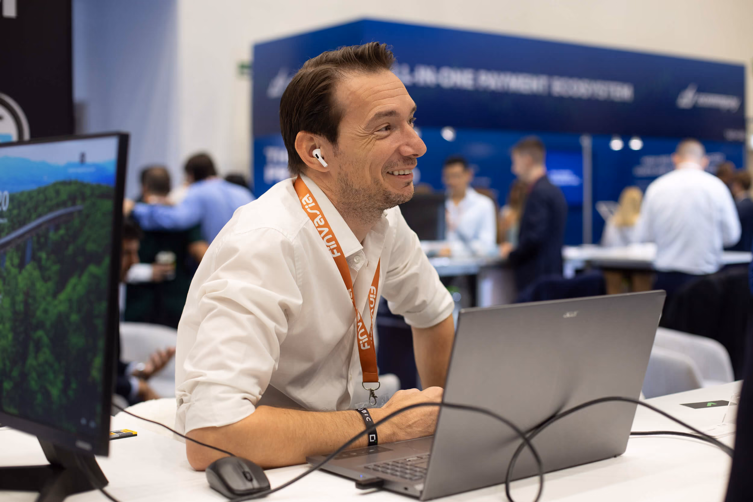 A smiling Match-Prime team member working on a laptop at an industry expo, the exhibition hall busy behind him.