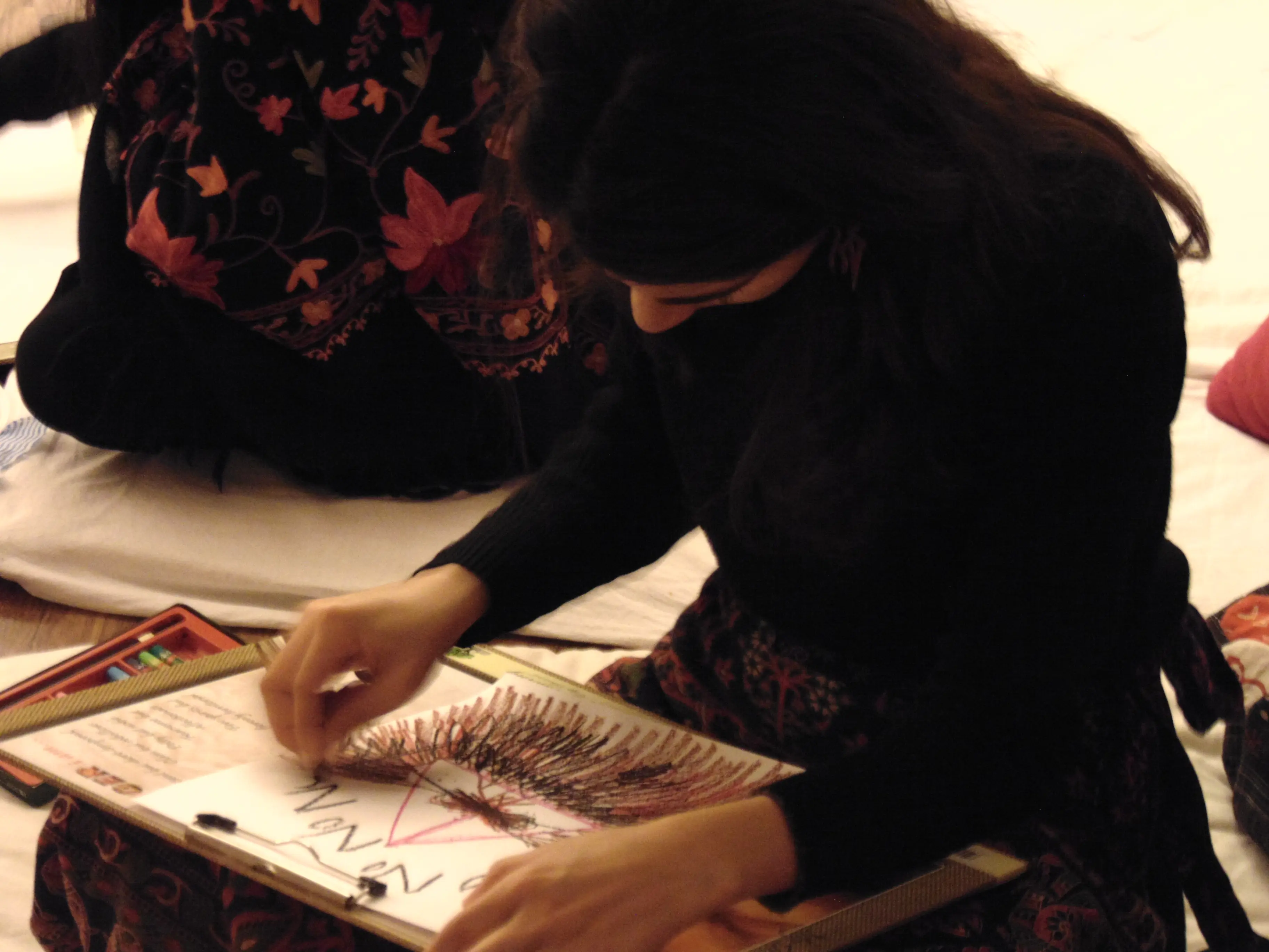 Woman in dark clothing seated, focused on creating colorful art with colored pencils on a sketchpad. Another person is partially visible beside her.