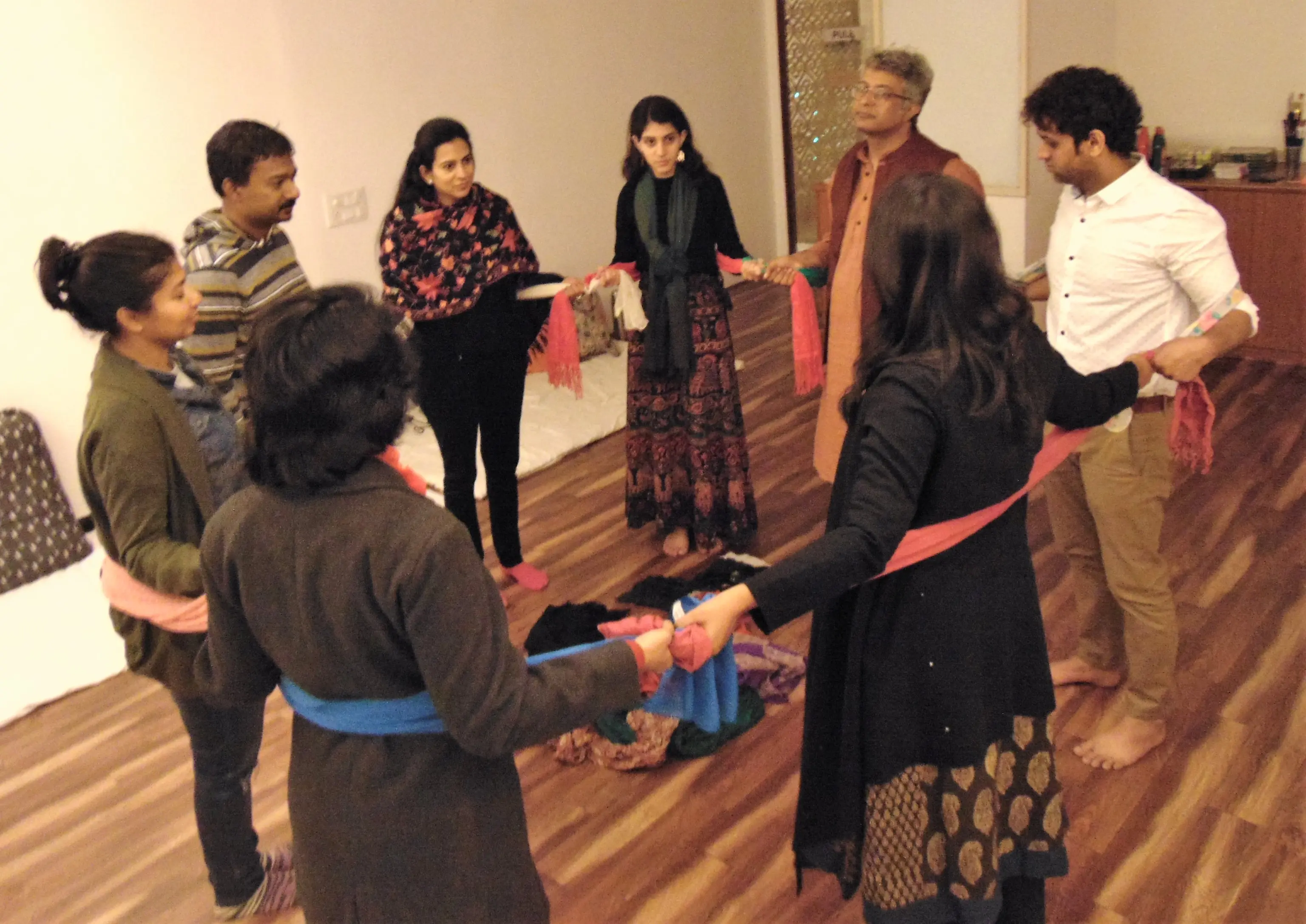 A group of eight people stand in a circle on a wooden floor, holding colorful scarves. The setting is calm and collaborative, suggesting a workshop.
