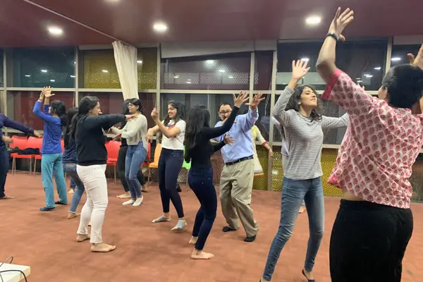 A group of people in casual attire energetically dance in a well-lit room with large windows. Their expressions convey joy and engagement, creating a lively atmosphere.