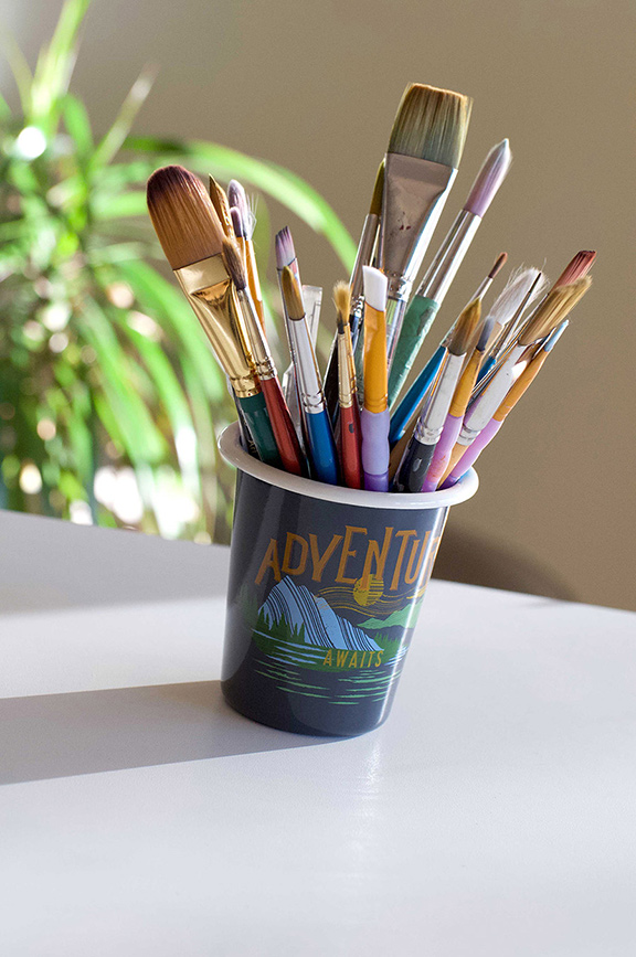 A dark cup holding various paintbrushes with colorful handles, displaying the text 'ADVENTURE AWAITS' with a mountain and sun illustration, set on a white surface with a blurred green plant in the background.
