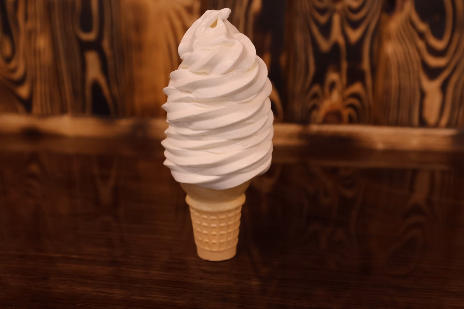 Soft serve vanilla ice cream cone with swirled peak on a wooden surface with a wood-paneled background.