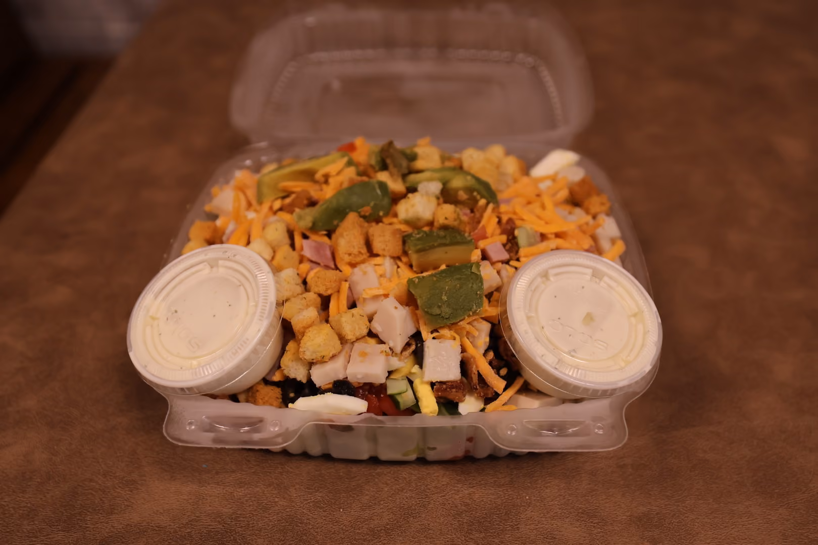 Takeout container with chopped salad including avocado, cheese, turkey, croutons, and two containers of dressing.