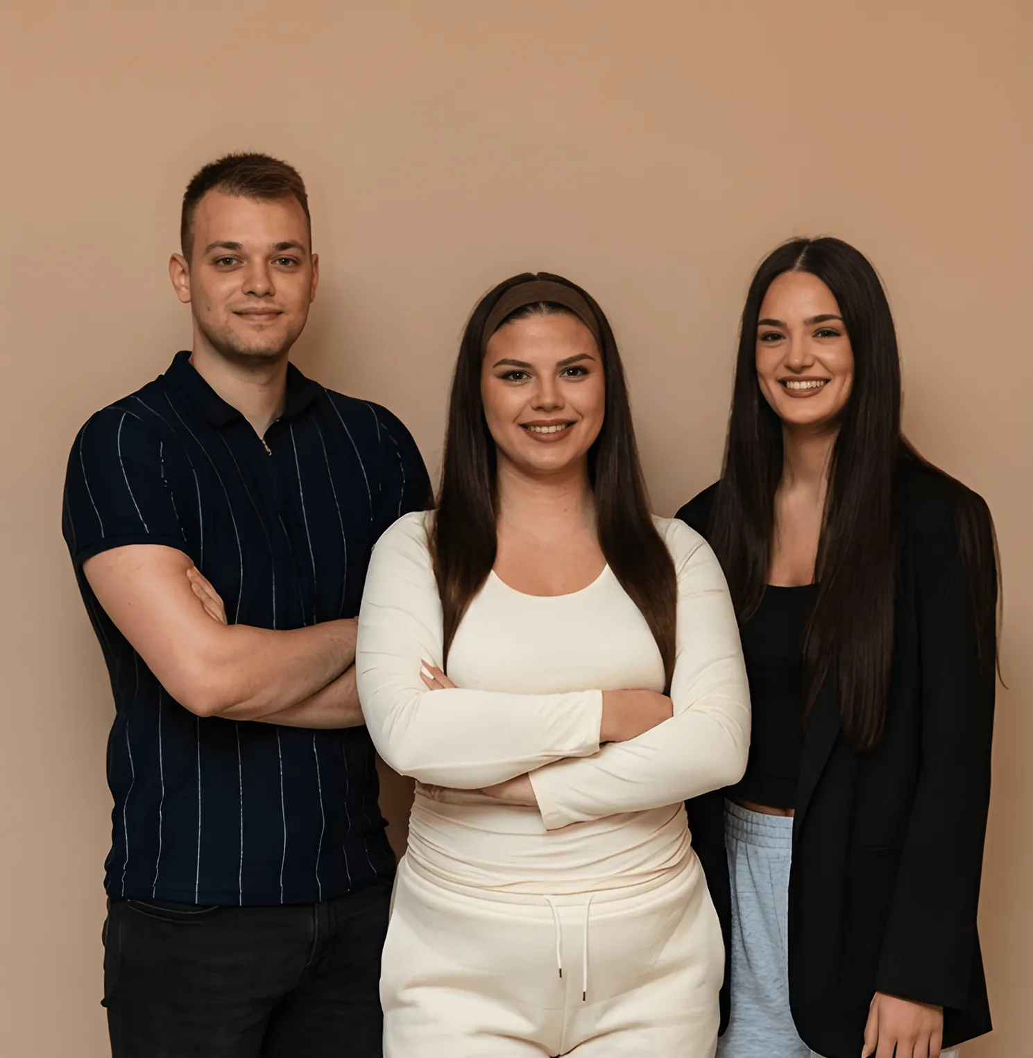 Three young adults standing side by side with arms crossed, smiling at the camera against a beige background.