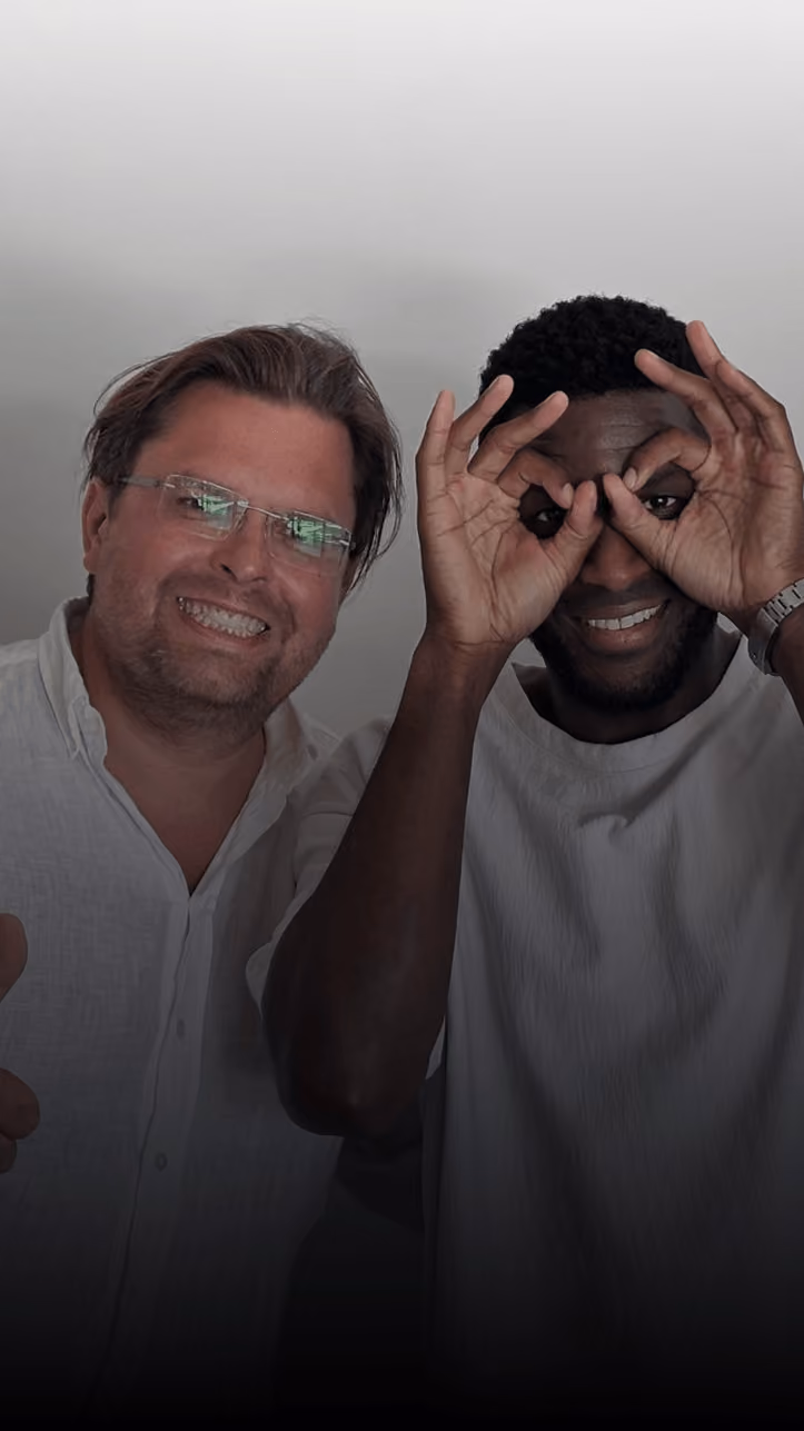 Zwei lächelnde Männer, einer zeigt eine Brille-Geste mit den Händen, beide tragen weiße Hemden vor neutralem Hintergrund.