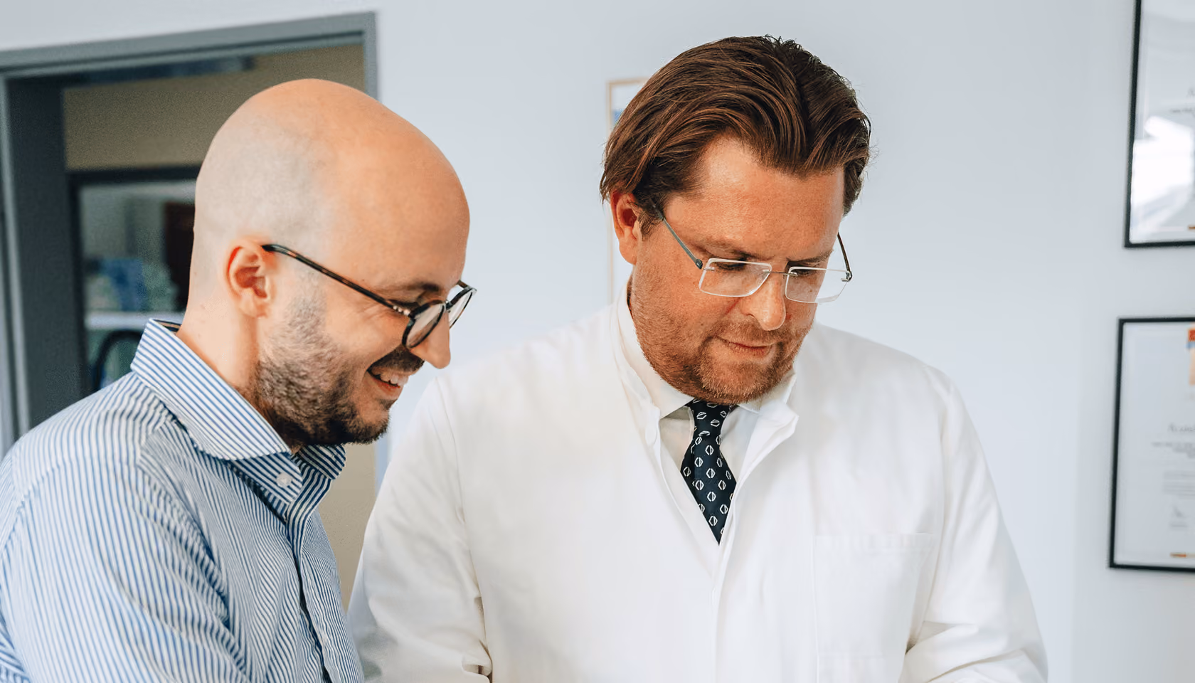 Zwei Männer im Gespräch, einer im Anzug, der andere in einem weißen Arztkittel, in einem Büro mit Zertifikaten an der Wand.