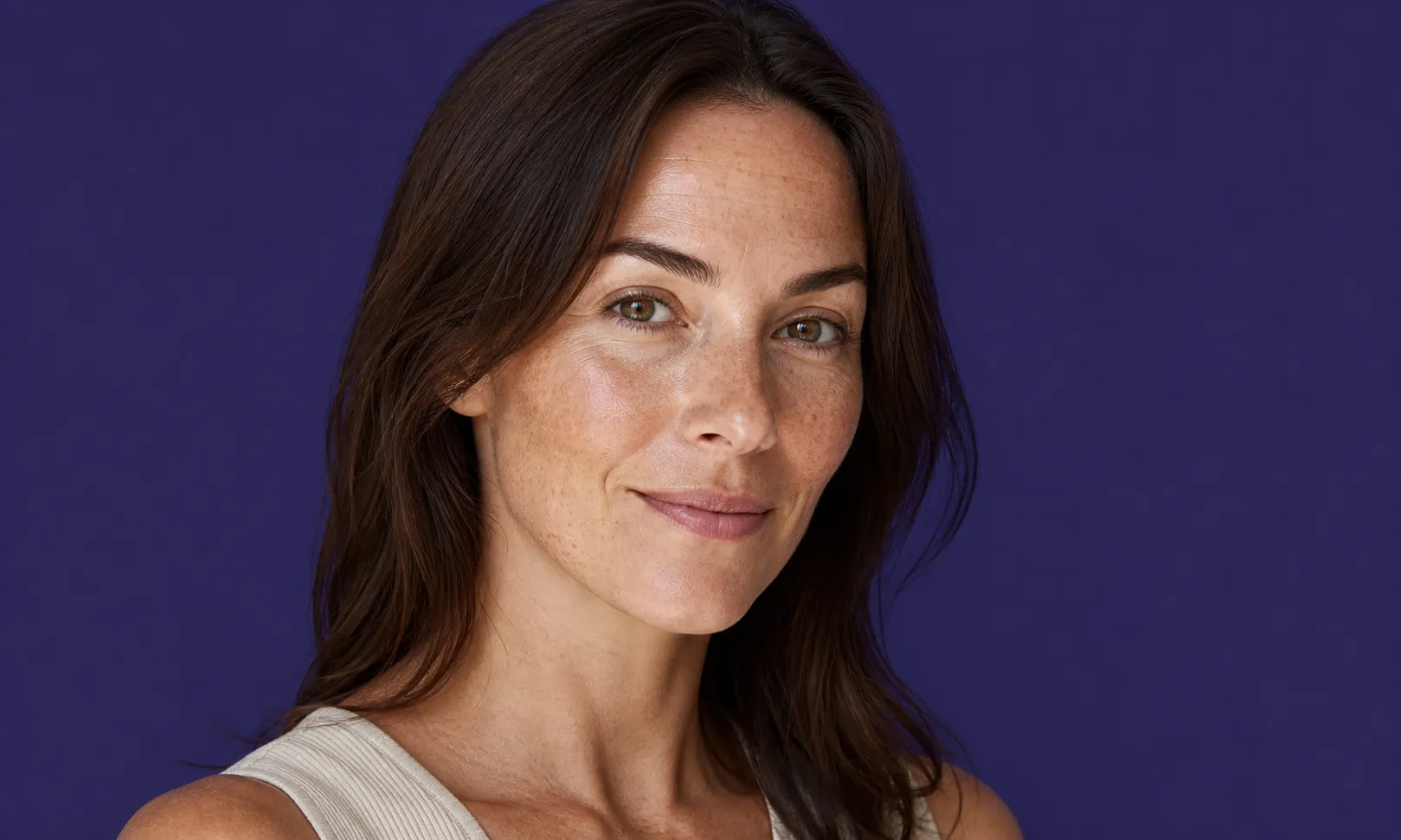 Femme souriante aux cheveux bruns et taches de rousseur sur fond violet.