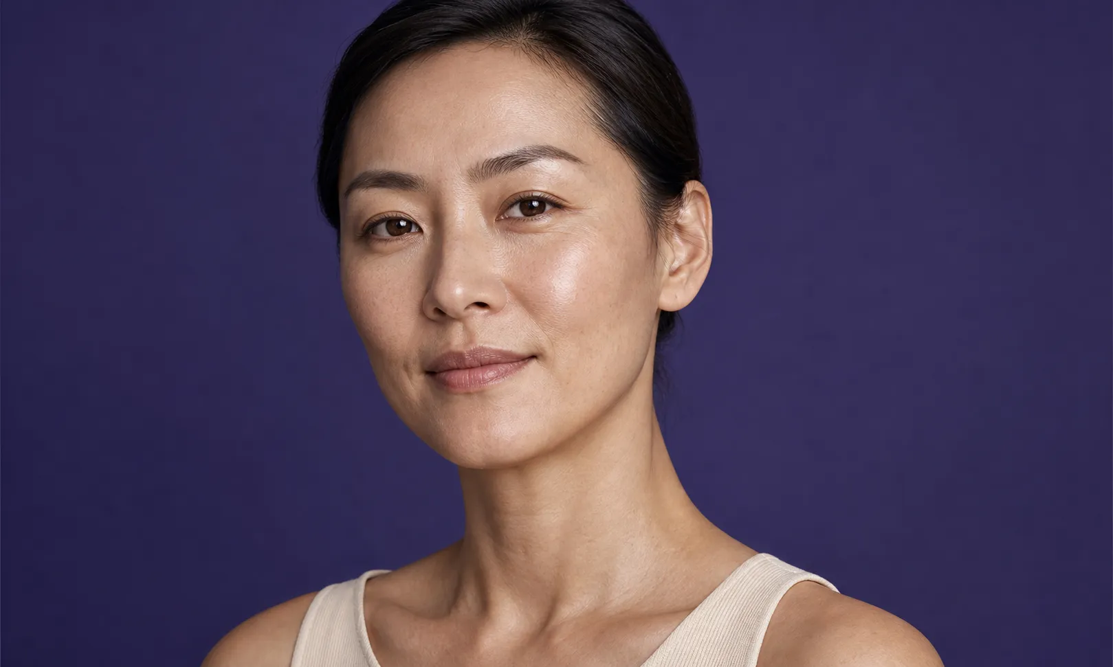 Femme asiatique au visage naturel souriant doucement sur fond violet uni.