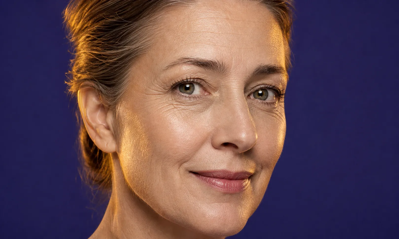 Gros plan d'une femme d'âge moyen avec peau claire, légères rides, souriant doucement sur fond bleu.