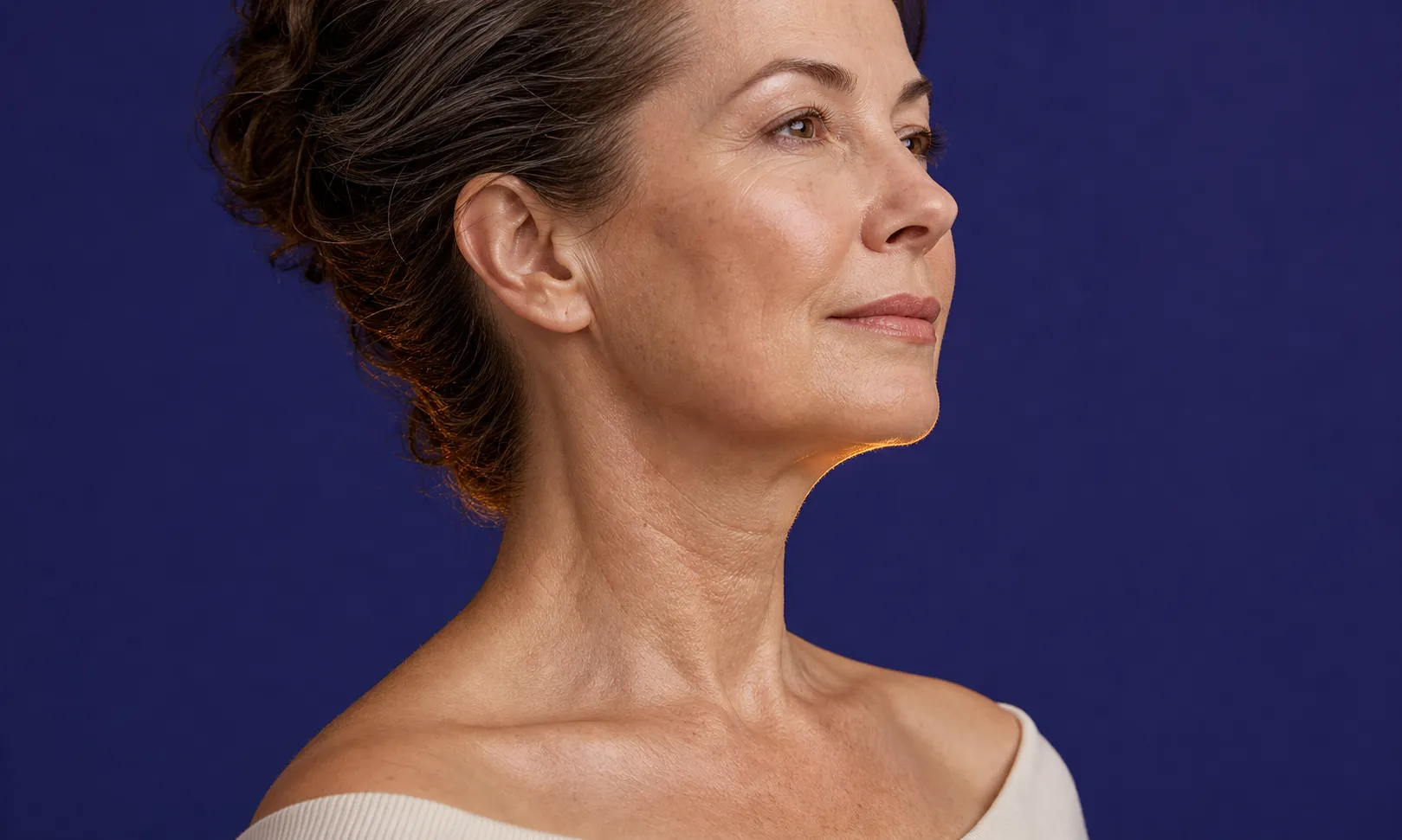 Portrait de profil d’une femme d’âge moyen avec peau lisse et regard confiant sur fond bleu.