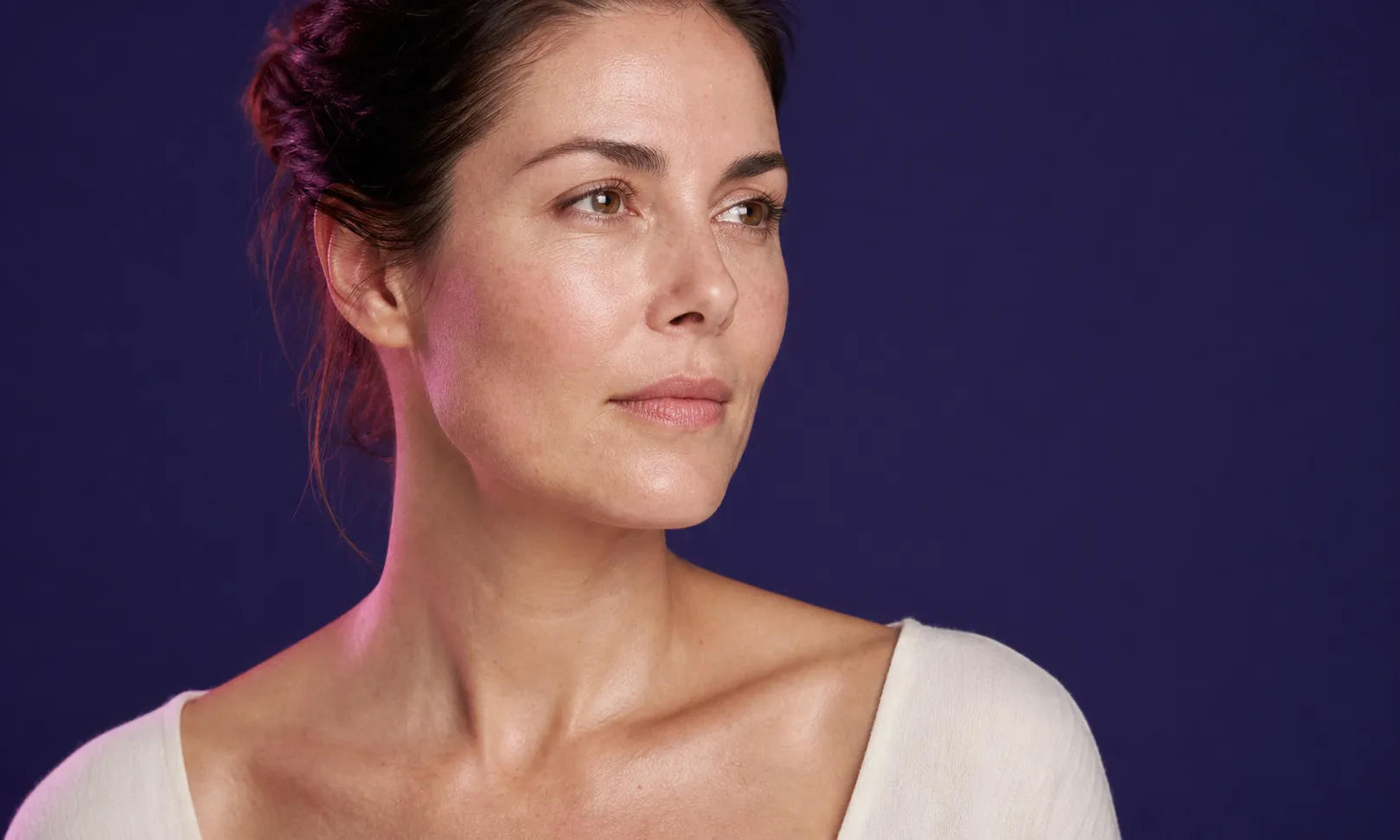Portrait d'une femme avec une peau éclatante regardant vers la droite sur fond violet foncé.