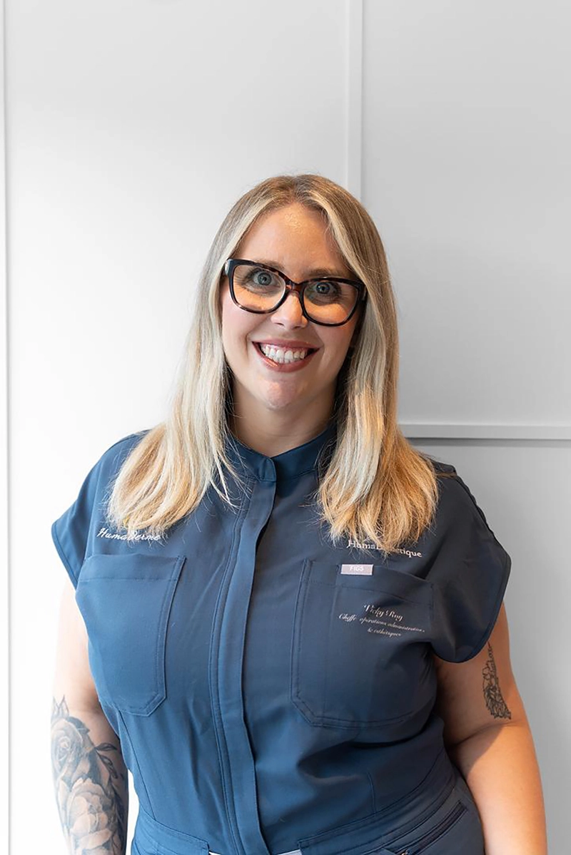 Une femme souriante avec des lunettes à monture noire, cheveux blonds mi-longs, portant une blouse bleue avec des poches sur le devant, et des tatouages visibles sur ses bras, posant devant un mur blanc simple.