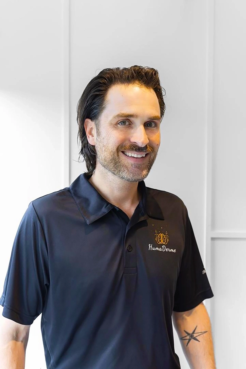 Homme souriant aux cheveux bruns mi-longs portant un polo noir avec le logo HumaDerme, avec un tatouage d'étoile sur l'avant-bras droit, sur fond blanc minimaliste.