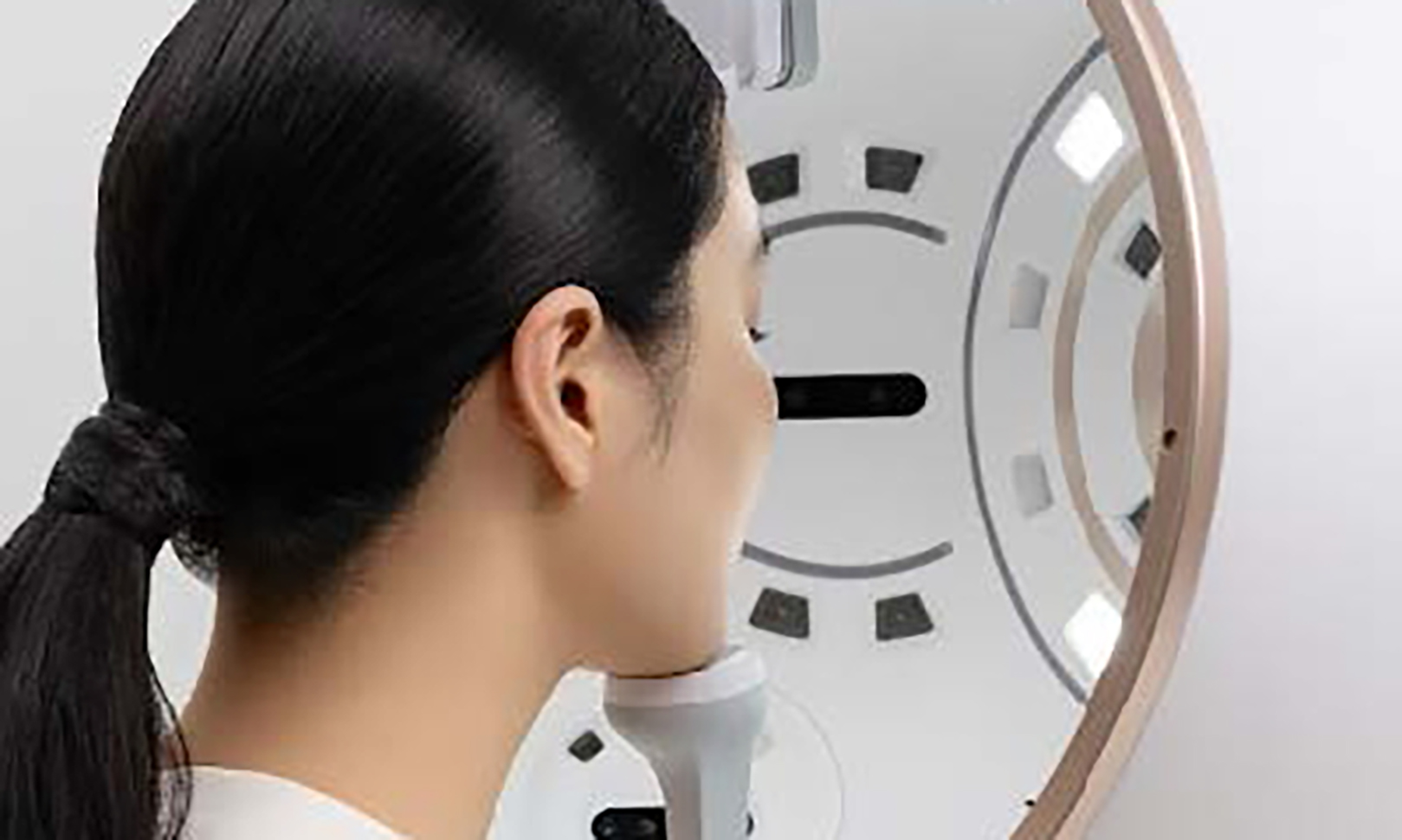 Femme vue de profil tenant un dispositif blanc près de son menton, devant un miroir rond avec des lumières intégrées et des repères noirs.