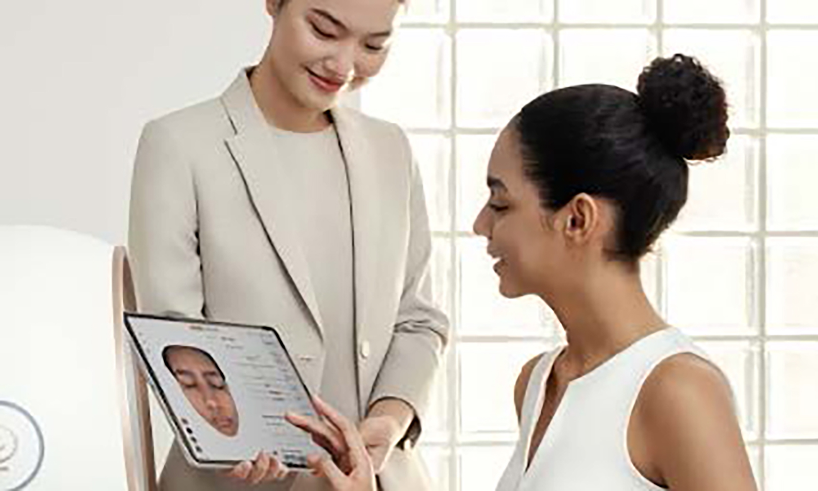 Deux femmes discutant, l'une en tailleur beige tenant une tablette affichant une analyse faciale, l'autre en haut blanc regardant l'écran et souriant.