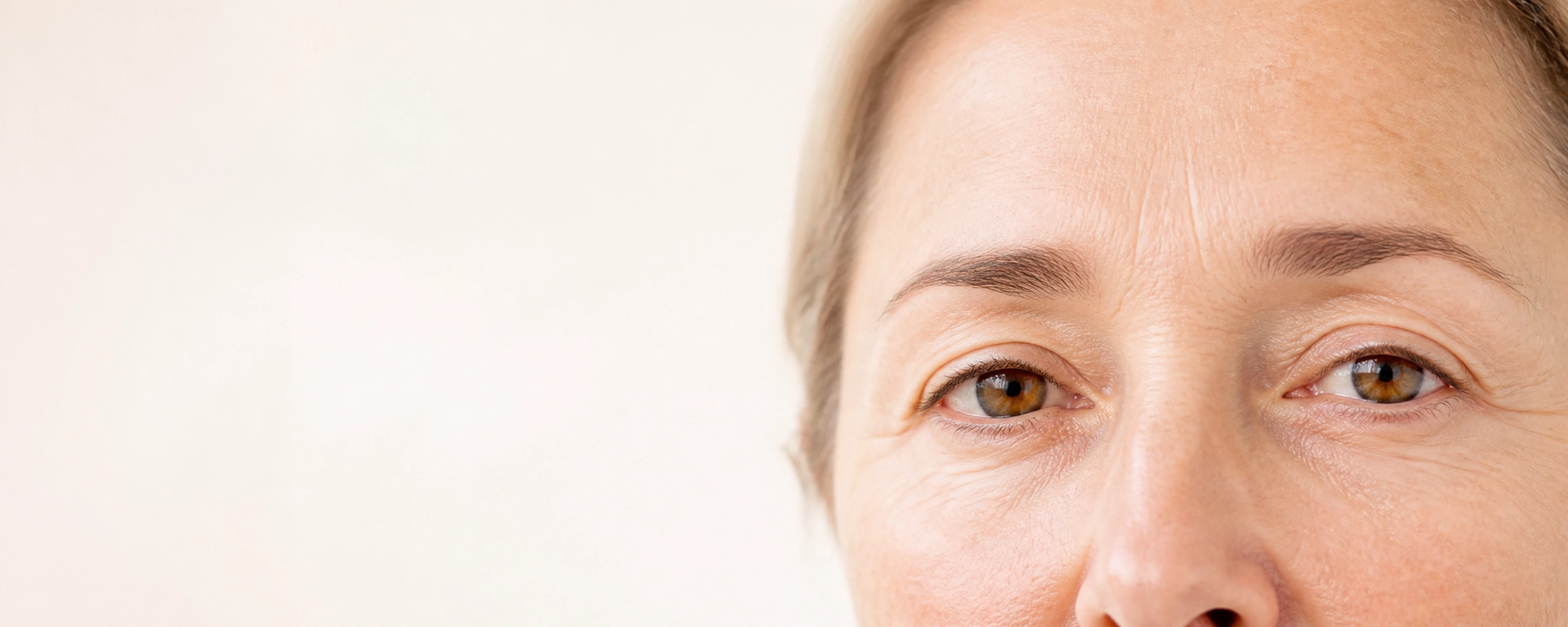 Vue rapprochée des yeux d'une personne d'âge moyen avec des yeux marron clair et des rides visibles sur le front et autour des yeux, sur un fond clair flou.
