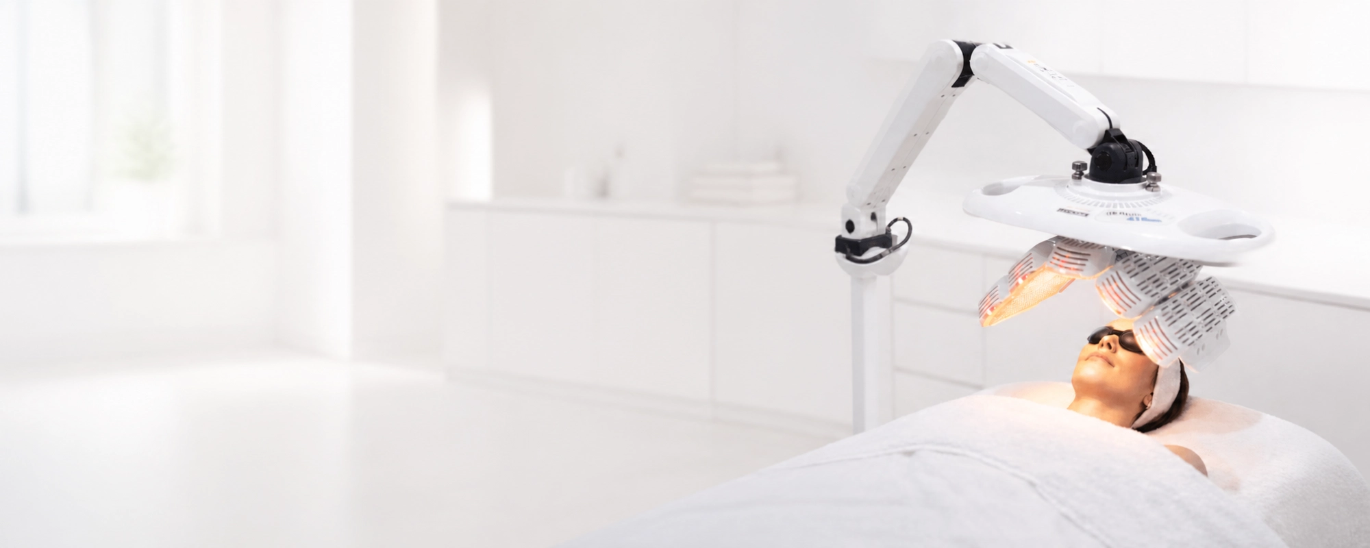 Femme allongée sous une lampe LED de luminothérapie sur un lit de spa, portant des lunettes de protection dans un environnement blanc et lumineux.