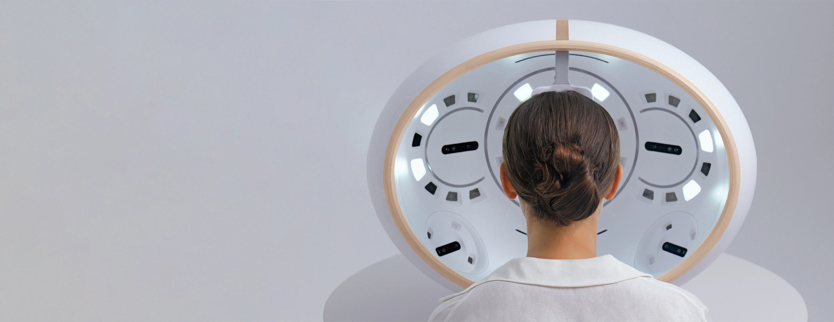 Vue arrière d'une femme avec les cheveux attachés en chignon regardant un miroir ovale éclairé avec des lumières blanches intégrées sur un fond gris clair.