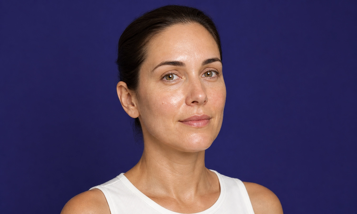 Portrait d'une femme souriante avec un teint éclatant et naturel, portant un débardeur blanc, sur un fond bleu uni.