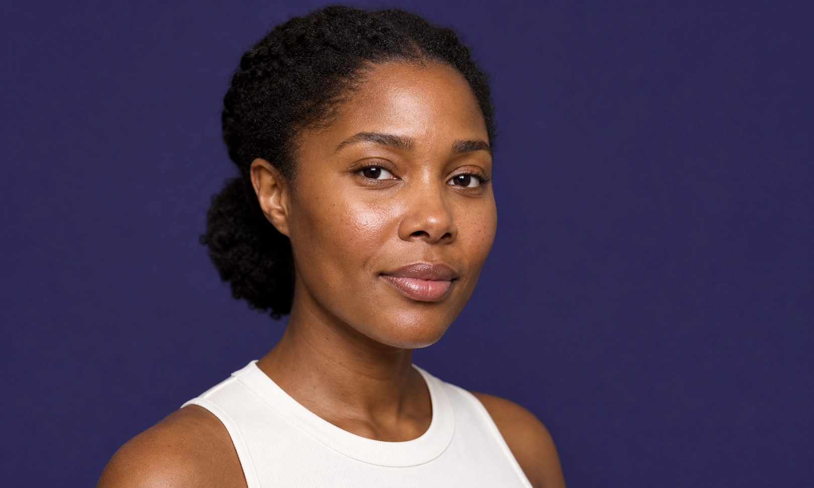 Portrait d'une femme noire avec les cheveux bouclés attachés, portant un débardeur blanc, sur fond violet.