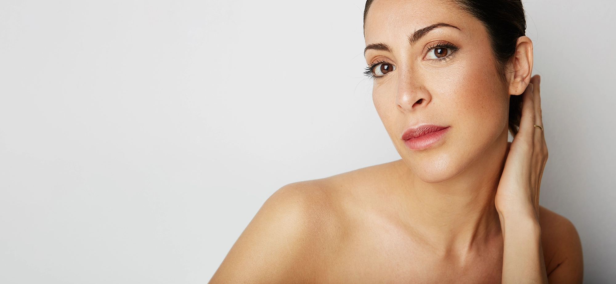 Femme avec une peau claire et un teint naturel, regardant vers l'avant avec une main touchant son cou, sur fond blanc.