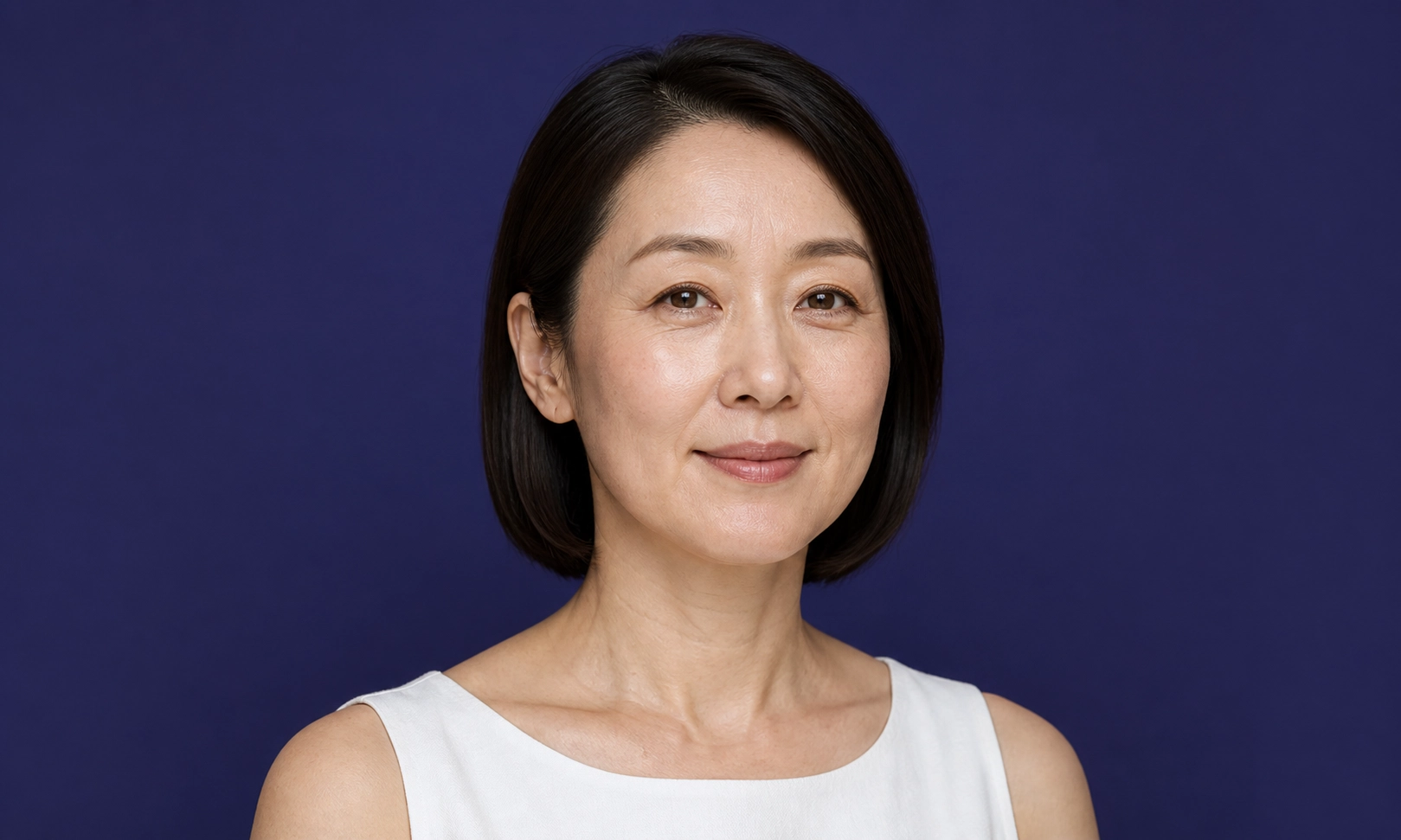 Portrait d'une femme asiatique d'âge moyen, aux cheveux noirs coupés au carré, portant un haut blanc sans manches, sur fond bleu marine.