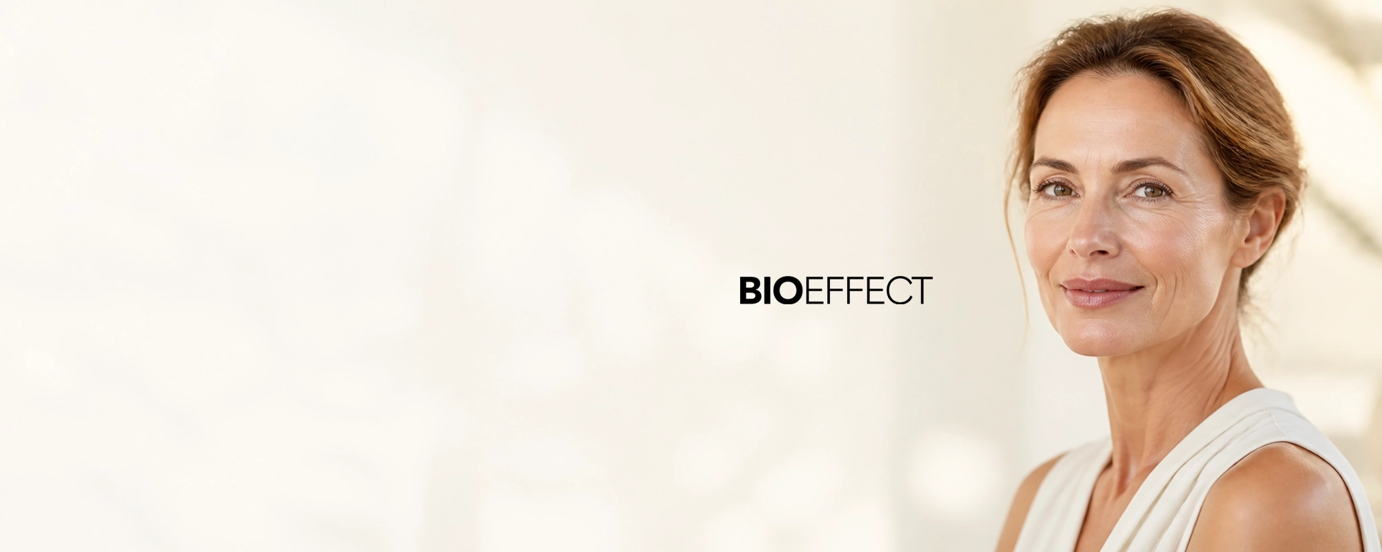 Femme d'âge moyen avec un teint lumineux et naturel, portant un haut sans manches clair, souriant doucement avec le logo BIOEFFECT à côté d'elle sur un fond clair.
