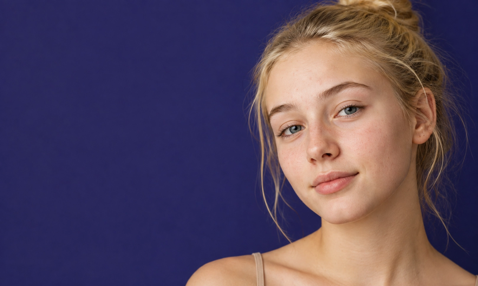 Jeune femme blonde aux yeux bleus avec la peau claire et cheveux attachés en chignon, posant devant un fond bleu uni.