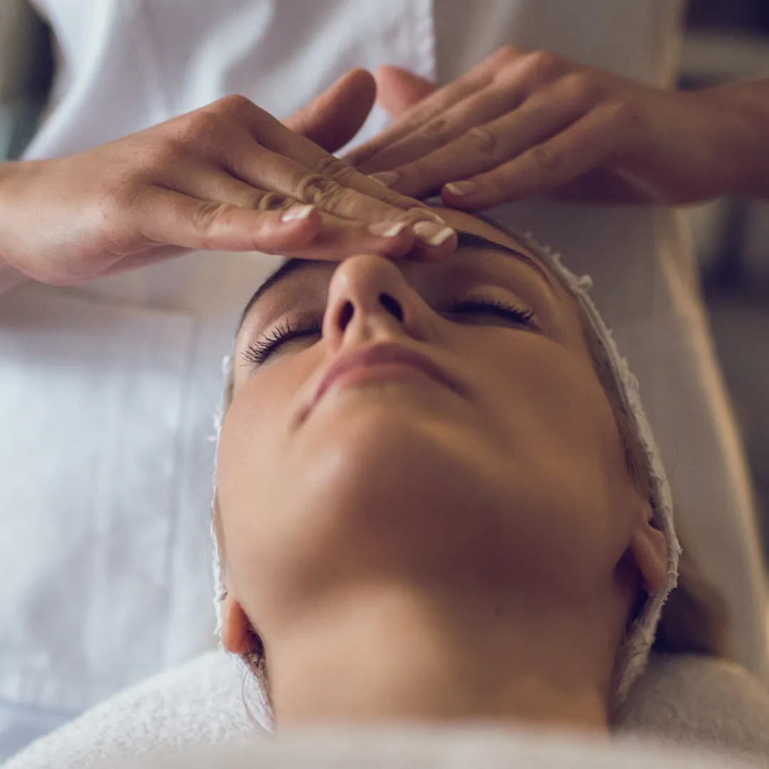 Une femme recevant un soin du visage, les yeux fermés, pendant qu'une personne masse doucement son front avec les mains.