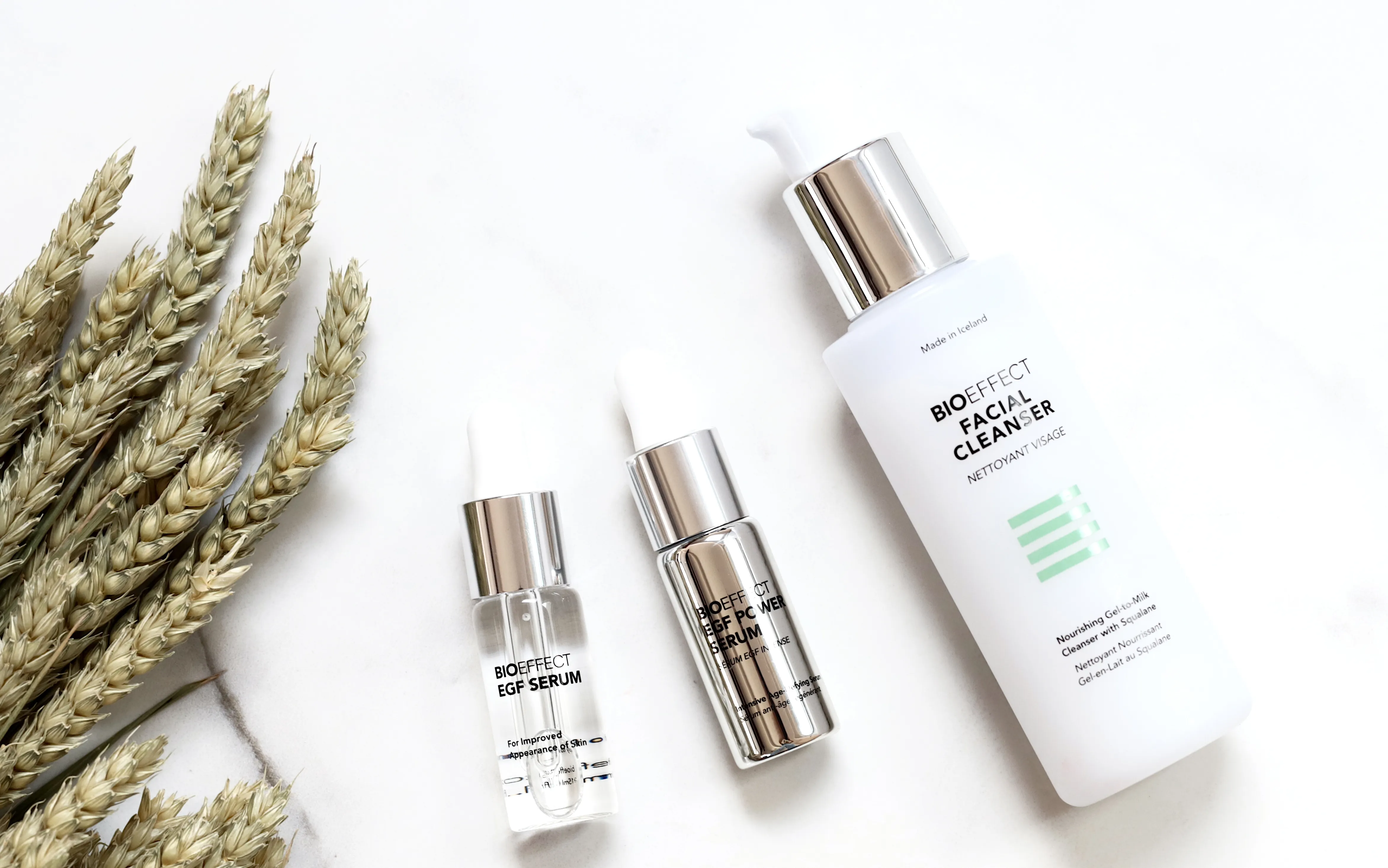 Trois produits de soin BIOEFFECT sur fond blanc, de gauche à droite : un petit flacon transparent avec pipette EGF Serum, un flacon argenté EGF Power Serum, et un flacon blanc avec pompe BIOEFFECT Facial Cleanser à base de gel-lait nourrissant et squalane, avec un bouquet de blé séché à gauche.
