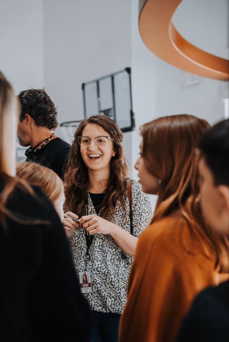 Smiling woman with glasses and ID badge talking with a group of people indoors.